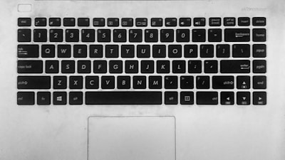 A monochrome image of a laptop keyboard featuring standard QWERTY layout. Keys include letters, numbers, and function keys as well as shortcuts such as Ctrl, Alt, and Windows key. The surface is slightly worn, indicating use. Below the keyboard, a touchpad is visible.