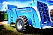 A large, blue agricultural machine with a prominent tire and multiple mechanical components is displayed outdoors on a grassy area. The machine is labeled with various markings, including 'Euromilk Buffalo RX900.' The backdrop includes other machinery and a partly cloudy sky.