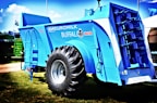 A large, blue agricultural machine with a prominent tire and multiple mechanical components is displayed outdoors on a grassy area. The machine is labeled with various markings, including 'Euromilk Buffalo RX900.' The backdrop includes other machinery and a partly cloudy sky.