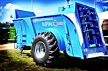 A large, blue agricultural machine with a prominent tire and multiple mechanical components is displayed outdoors on a grassy area. The machine is labeled with various markings, including 'Euromilk Buffalo RX900.' The backdrop includes other machinery and a partly cloudy sky.