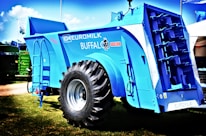 A large, blue agricultural machine with a prominent tire and multiple mechanical components is displayed outdoors on a grassy area. The machine is labeled with various markings, including 'Euromilk Buffalo RX900.' The backdrop includes other machinery and a partly cloudy sky.