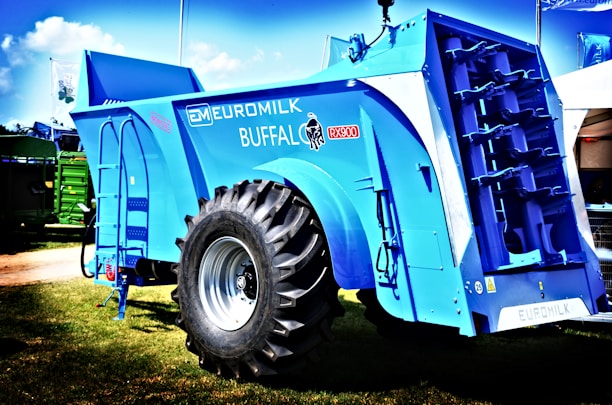 A large, blue agricultural machine with a prominent tire and multiple mechanical components is displayed outdoors on a grassy area. The machine is labeled with various markings, including 'Euromilk Buffalo RX900.' The backdrop includes other machinery and a partly cloudy sky.