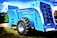A large, blue agricultural machine with a prominent tire and multiple mechanical components is displayed outdoors on a grassy area. The machine is labeled with various markings, including 'Euromilk Buffalo RX900.' The backdrop includes other machinery and a partly cloudy sky.