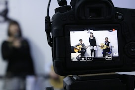 A camera is focused on a scene where a woman holds a microphone between two men playing guitars. The camera's LCD screen displays this scene, showing the details of the instruments and the expressions of the performers.
