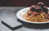 A plate of spaghetti topped with meatballs and tomato sauce is placed next to a smartphone on a dark surface. The food is presented on a white plate, and the lighting creates a cozy and inviting atmosphere.