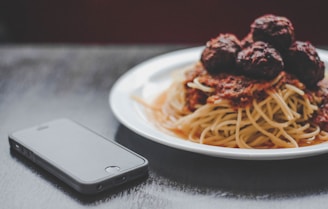 A plate of spaghetti topped with meatballs and tomato sauce is placed next to a smartphone on a dark surface. The food is presented on a white plate, and the lighting creates a cozy and inviting atmosphere.
