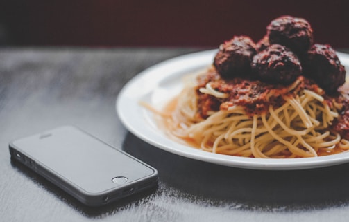 A plate of spaghetti topped with meatballs and tomato sauce is placed next to a smartphone on a dark surface. The food is presented on a white plate, and the lighting creates a cozy and inviting atmosphere.