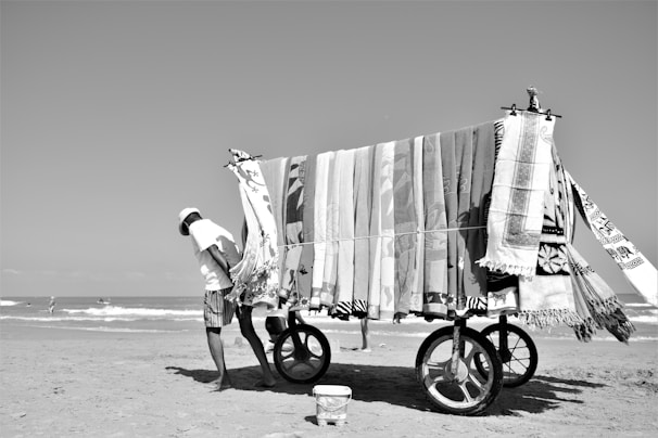 A person pushing a cart loaded with various patterned towels on a sandy beach. The sky is clear, and there are waves in the background with some people enjoying the water.
