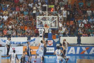 A basketball court scene with players jumping for a rebound and a live score display overhead.