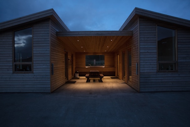 A modern wooden building with a sheltered central seating area, illuminated by recessed lights, creating a warm contrast against the evening sky. Large windows frame the structure, hinting at a minimalist and contemporary design style.