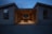A modern wooden building with a sheltered central seating area, illuminated by recessed lights, creating a warm contrast against the evening sky. Large windows frame the structure, hinting at a minimalist and contemporary design style.