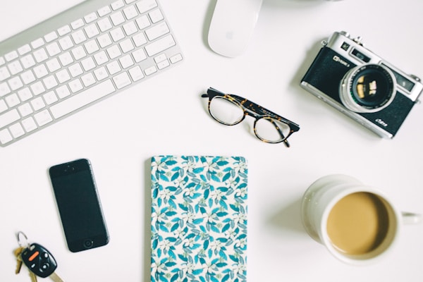 A neatly arranged desk layout featuring a white keyboard, a wireless mouse, a vintage camera, a pair of eyeglasses, a smartphone, a set of car keys, a notebook with a blue floral cover, and a mug filled with coffee. The items are placed on a white surface, giving a minimalistic and organized look.