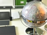 A close-up view of a silver metallic globe placed on a desk, with visible geographical markings and names. The background includes blurred office items such as a keyboard and a tablet.