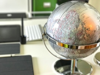 A close-up view of a silver metallic globe placed on a desk, with visible geographical markings and names. The background includes blurred office items such as a keyboard and a tablet.