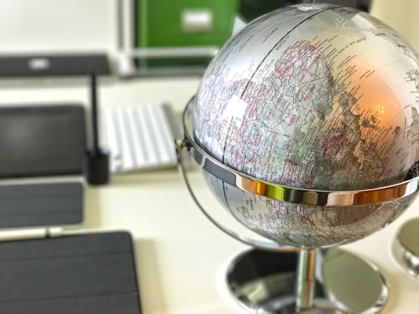 A close-up view of a silver metallic globe placed on a desk, with visible geographical markings and names. The background includes blurred office items such as a keyboard and a tablet.