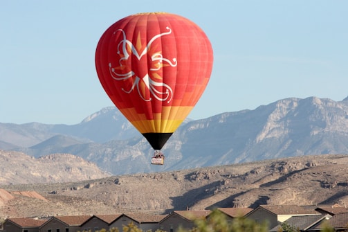 A red hot air balloon with intricate white patterns and a yellow band near the bottom floats above a residential area. The background features a rugged mountain range under a clear blue sky.