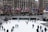 A number of people are ice skating at an outdoor rink in a city setting. The rink is surrounded by a barrier, and flags of various countries are lined up above the rink. Behind the rink are buildings with tall windows and leafless trees. There are also some structures resembling restaurants or shops adjacent to the rink.