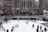 A number of people are ice skating at an outdoor rink in a city setting. The rink is surrounded by a barrier, and flags of various countries are lined up above the rink. Behind the rink are buildings with tall windows and leafless trees. There are also some structures resembling restaurants or shops adjacent to the rink.