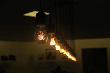 A row of glowing incandescent light bulbs hanging from the ceiling in a dimly lit room, creating a warm ambiance. The focus is on the closest bulb, with others gradually fading into the background.