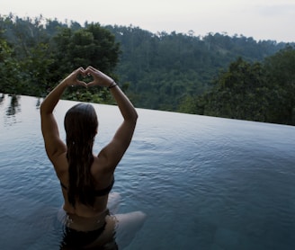 woman in infinity pool making heart hand gesture facing green leafed trees