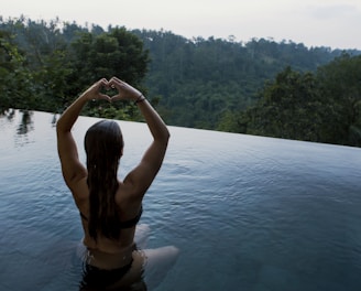 woman in infinity pool making heart hand gesture facing green leafed trees