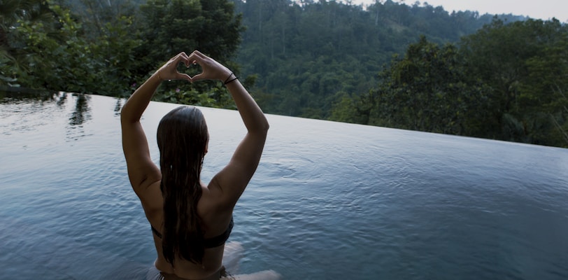 woman in infinity pool making heart hand gesture facing green leafed trees