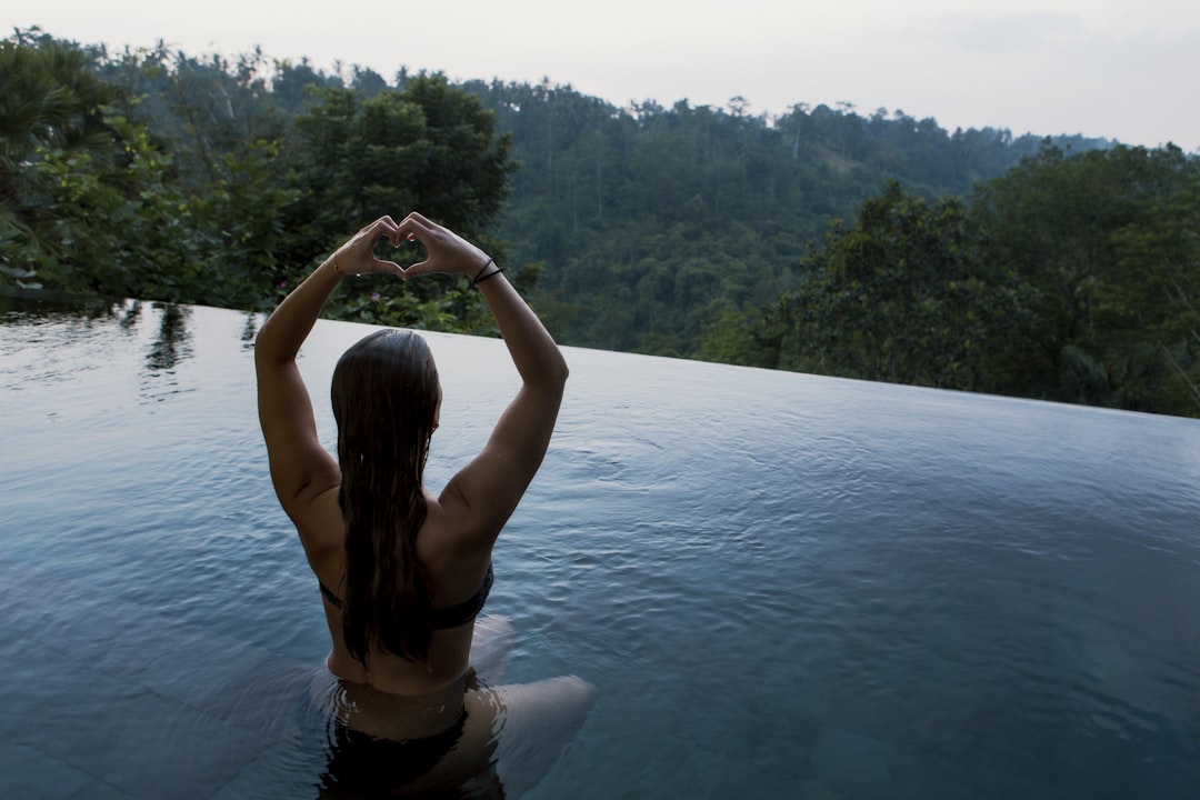 woman in infinity pool making heart hand gesture facing green leafed trees, pool meditation