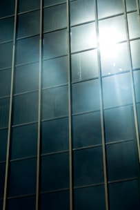 Tall glass windows reflect sunlight, creating a bright and glaring effect across the surface. The light bounces off the glass, casting beams and a soft glare in the surrounding areas.