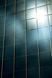 Tall glass windows reflect sunlight, creating a bright and glaring effect across the surface. The light bounces off the glass, casting beams and a soft glare in the surrounding areas.