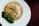A dish featuring a mixture of mushrooms, carrots, and possibly other ingredients wrapped in a thin pastry or tofu skin, served in a light broth. Broccoli florets are placed on the side as a garnish on a white plate against a dark background.