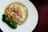 A dish featuring a mixture of mushrooms, carrots, and possibly other ingredients wrapped in a thin pastry or tofu skin, served in a light broth. Broccoli florets are placed on the side as a garnish on a white plate against a dark background.