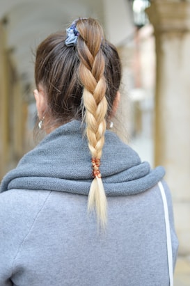 A person wearing a gray sweater has their hair styled in a braided ponytail. The hair is secured with a blue scrunchie at the top and decorated with small rings towards the end. The background is a softly blurred urban setting.