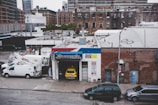 An urban scene featuring a garage named Mike's Auto Electrics with a yellow car parked inside. Several white utility trucks are parked nearby, and the backdrop consists of brick buildings and city structures. The setting suggests an industrial or repair district within a city.