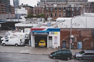 An urban scene featuring a garage named Mike's Auto Electrics with a yellow car parked inside. Several white utility trucks are parked nearby, and the backdrop consists of brick buildings and city structures. The setting suggests an industrial or repair district within a city.