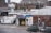An urban scene featuring a garage named Mike's Auto Electrics with a yellow car parked inside. Several white utility trucks are parked nearby, and the backdrop consists of brick buildings and city structures. The setting suggests an industrial or repair district within a city.