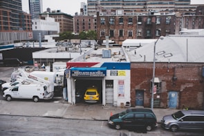An urban scene featuring a garage named Mike's Auto Electrics with a yellow car parked inside. Several white utility trucks are parked nearby, and the backdrop consists of brick buildings and city structures. The setting suggests an industrial or repair district within a city.