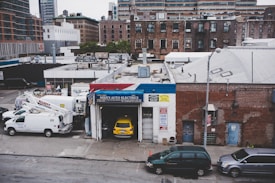 An urban scene featuring a garage named Mike's Auto Electrics with a yellow car parked inside. Several white utility trucks are parked nearby, and the backdrop consists of brick buildings and city structures. The setting suggests an industrial or repair district within a city.