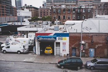 An urban scene featuring a garage named Mike's Auto Electrics with a yellow car parked inside. Several white utility trucks are parked nearby, and the backdrop consists of brick buildings and city structures. The setting suggests an industrial or repair district within a city.