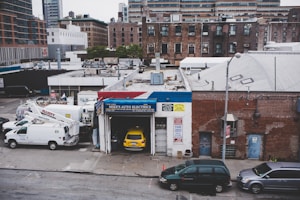 An urban scene featuring a garage named Mike's Auto Electrics with a yellow car parked inside. Several white utility trucks are parked nearby, and the backdrop consists of brick buildings and city structures. The setting suggests an industrial or repair district within a city.