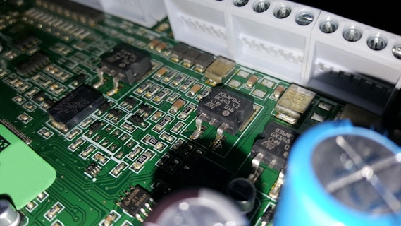 A close-up of a printed circuit board featuring various electronic components including integrated circuits, capacitors, and resistors. The green board is densely packed with intricate patterns and metallic connections. Some components have small markings, indicating their specifications.