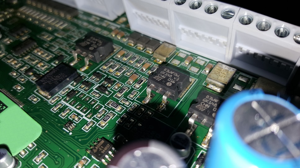 A close-up of a printed circuit board featuring various electronic components including integrated circuits, capacitors, and resistors. The green board is densely packed with intricate patterns and metallic connections. Some components have small markings, indicating their specifications.