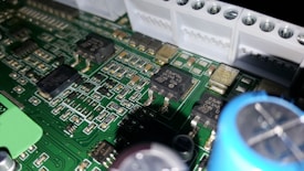 A close-up of a printed circuit board featuring various electronic components including integrated circuits, capacitors, and resistors. The green board is densely packed with intricate patterns and metallic connections. Some components have small markings, indicating their specifications.