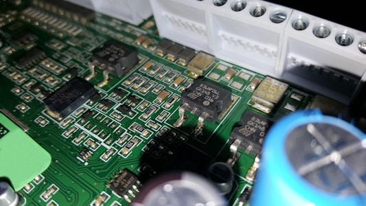 A close-up of a printed circuit board featuring various electronic components including integrated circuits, capacitors, and resistors. The green board is densely packed with intricate patterns and metallic connections. Some components have small markings, indicating their specifications.