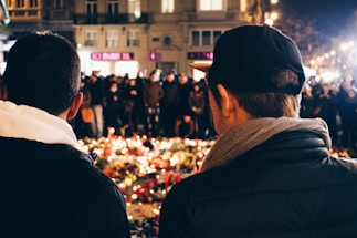 Two people are seen from the back, standing in front of a candlelit vigil. A crowd is gathered in the background, creating a solemn and respectful atmosphere. The scene takes place in an urban setting at night, with buildings softly illuminated.