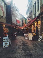 A narrow cobblestone street lined with eateries featuring colorful awnings and outdoor seating. Menus and signs are prominently displayed outside each establishment, indicating various dining options. The street is flanked by tall, narrow buildings with a mix of architectural styles.
