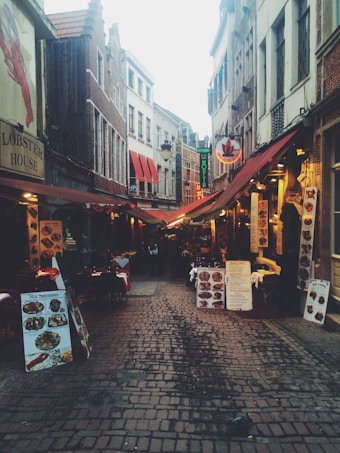 A narrow cobblestone street lined with eateries featuring colorful awnings and outdoor seating. Menus and signs are prominently displayed outside each establishment, indicating various dining options. The street is flanked by tall, narrow buildings with a mix of architectural styles.