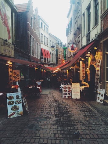 A narrow cobblestone street lined with eateries featuring colorful awnings and outdoor seating. Menus and signs are prominently displayed outside each establishment, indicating various dining options. The street is flanked by tall, narrow buildings with a mix of architectural styles.
