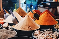 An assortment of colorful spices are displayed in conical piles at a market stall, each with a scoop. The spices vary in color, from bright oranges and yellows to deep browns. Various other spices and roots are visible in the foreground.