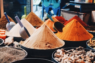 An assortment of colorful spices are displayed in conical piles at a market stall, each with a scoop. The spices vary in color, from bright oranges and yellows to deep browns. Various other spices and roots are visible in the foreground.
