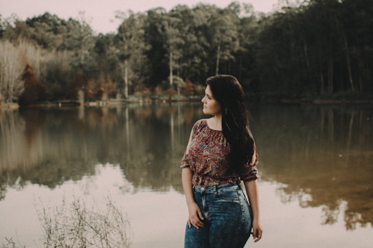 A woman stands near a calm lake, looking to the side. The background features dense trees and their reflections on the water. She is wearing a colorful floral top and jeans, with her long hair flowing over her shoulders. The atmosphere is serene and natural.